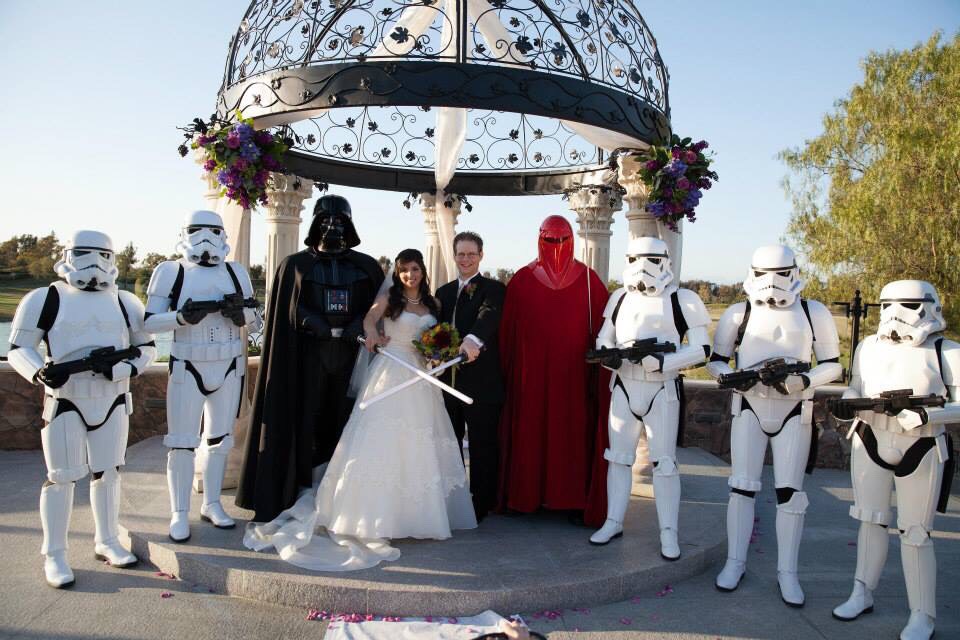 A newlywed couple smiles into the camera with their wedding party of stormtroopers, an Imperial guard, and Darth Vader after their Star Wars wedding