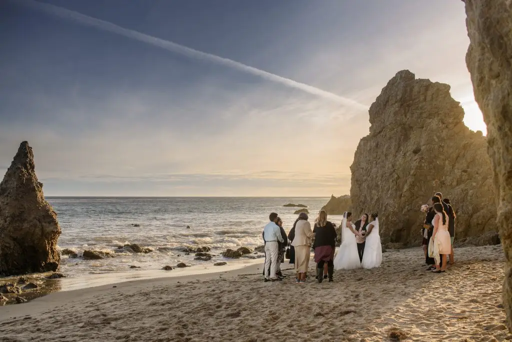 Two brides in white dresses hold hands while Let's Get Married by Marie conducts their ceremony with their loved ones on the beach as the sun sets behind them