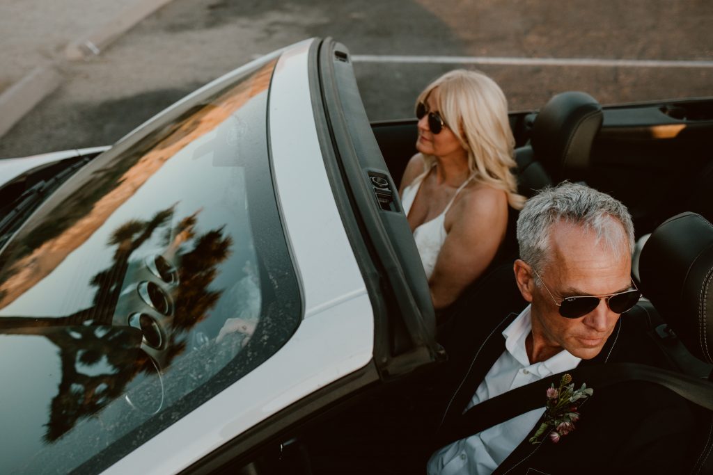 Cool newlyweds in their wedding clothes drive in a topless white convertible after eloping in Joshua tree