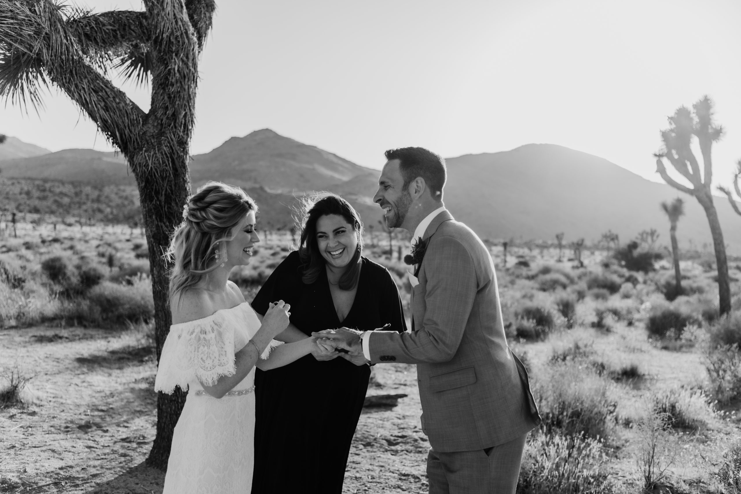 Let's Get Married by Marie laughs while performing a Joshua Tree elopement package for a smiling bride in a white boho dress and a laughing groom in a gray suit in Joshua Tree National Park