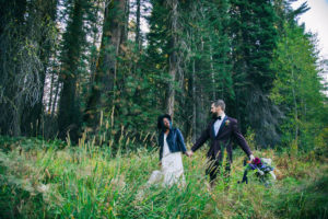 A black bride in a white dress and black leather jacket smirks at her new husband in a dark suit in a meadow during their Epic Elopement Experience for their Portland Elopement Packages