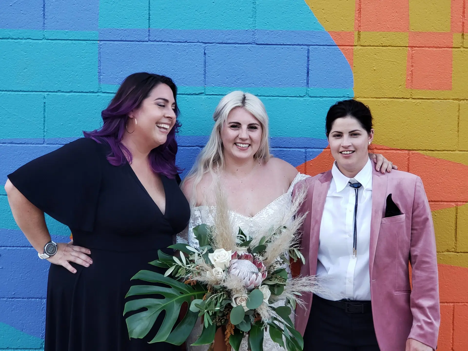 A bride in a white dress with a large tropical bouquet and a bride in a pink velvet jacket and white shirt smile into the camera with Let's Get Married by Marie Palm Springs elopement package