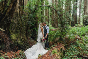 Redwood elopement packages by Let's Get Married by Marie show a black bride in a white wedding dress holds a bouquet and snuggles with her white groom in a suit during their Redwood elopement package with Let's Get Married by Marie