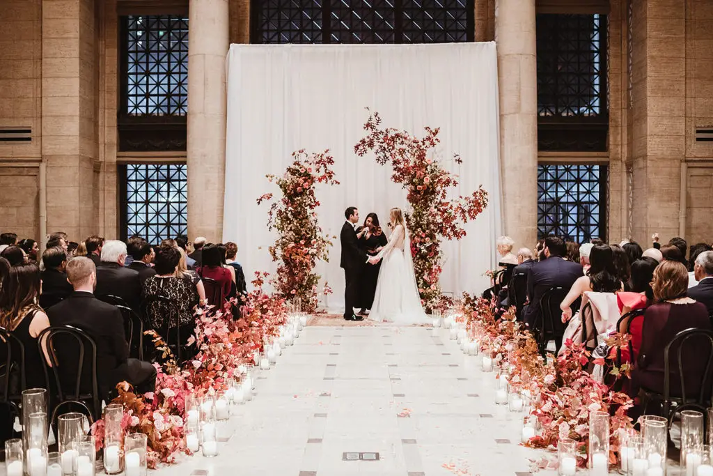 Minister Marie of Let's Get Married by Marie, a San Francisco wedding officiant, leads a wedding ceremony for a bride and groom surrounded by pink florals at the San Francisco Asian Art Museum