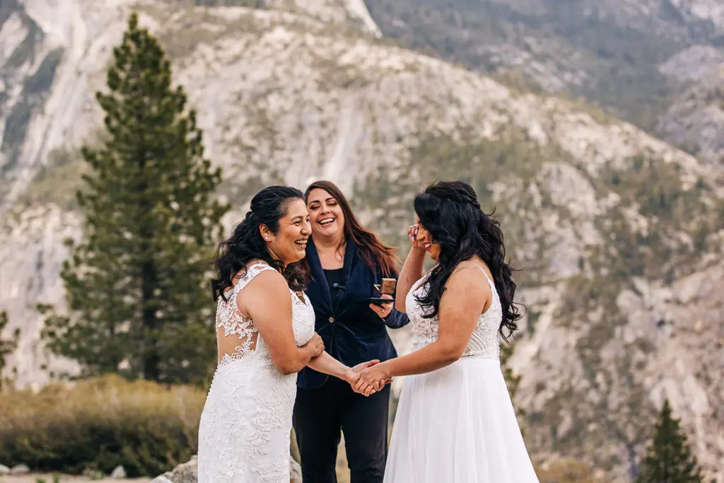 Let's Get Married by Marie officiates an elopement in Yosemite for two dark-haired brides in white wedding dresses with Half Dome in the background from their Yosemite elopement package location of Glacier Point