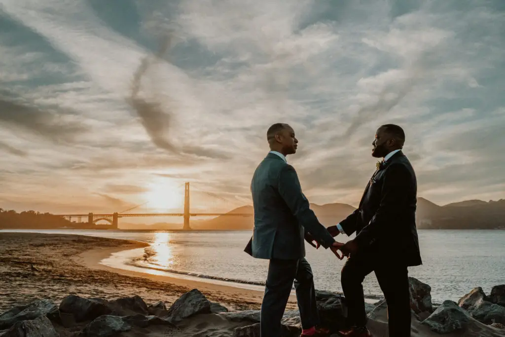 Two newlywed grooms hold hands in the foreground with the sun setting behind the Golden Gate bridge during their San Francisco elopement package