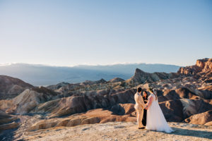 A groom in a tan suit and a bride in a white boho wedding dress stand elope in Death Valley with the sun setting behind them as Let's Get Married by Marie officiates their marriage ceremony