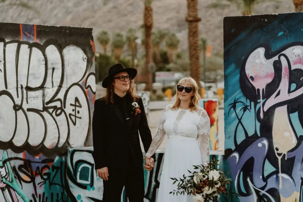 Palm Springs elopement packages with Let's Get Married by Marie featuring a groom wearing all black including a hat and sunglasses and a bride in a modern lace wedding dress and sunglasses in downtown Palm Springs